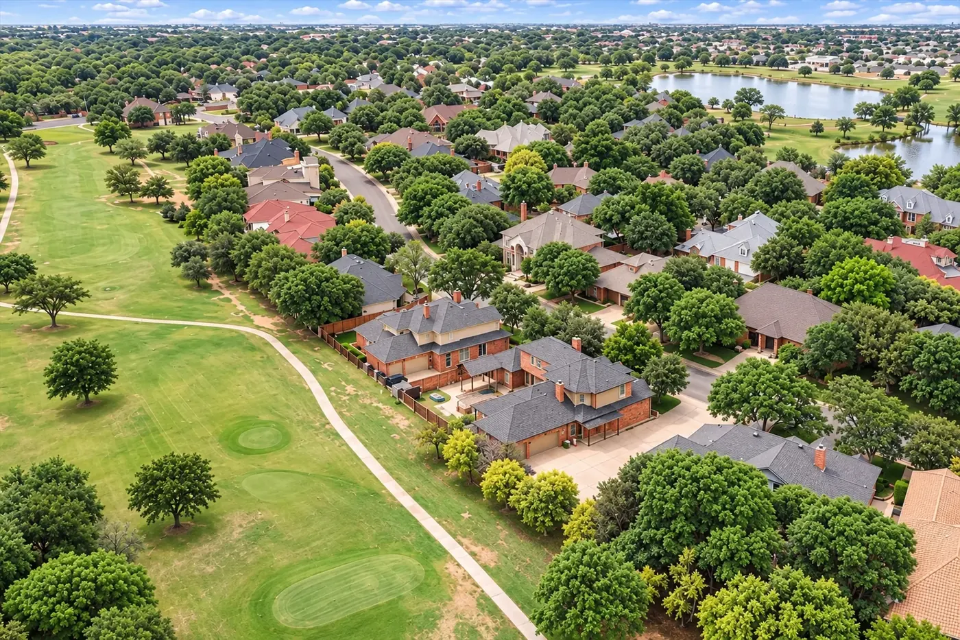 An aerial view of a golf course beside tree-lined streets of large homes, with a lake beyond.